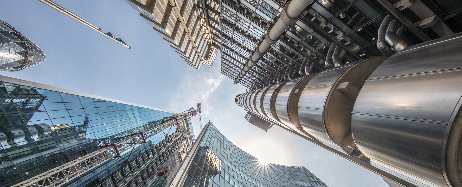 Looking directly up at the Lloyd's building and surrounding buildings to a clear, blue sky, the sun is shining off one of the buildings